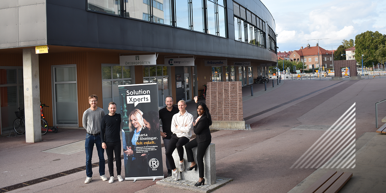 Kollegorna på Solution Xperts står med en roll-up utanför Örebrokontoret. En av medarbetarna pekar mot byggnaden.