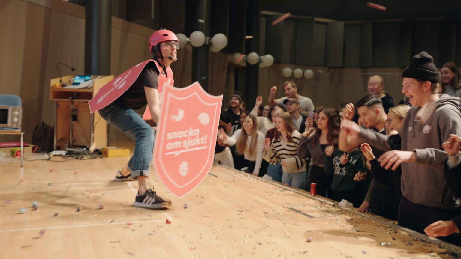 Bild från snacka om sjukt event. Person på scen med rosa hjälm och skylt som det står "Snacka om sjukt". Ungdomar i publiken som är glada och ser engagerade ut.